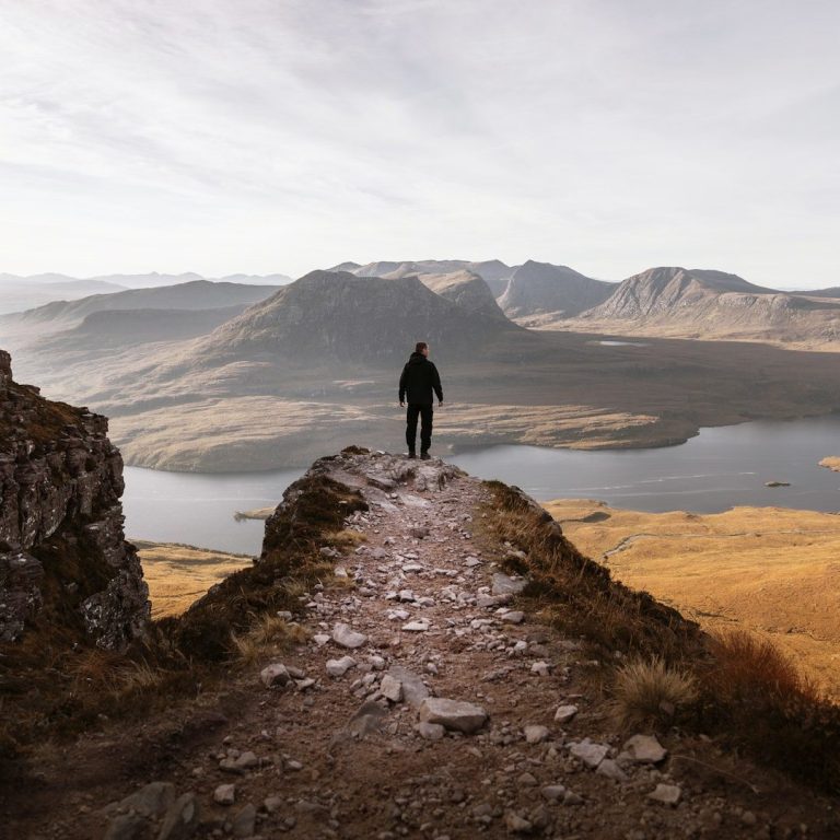 Ein Mann steht auf einem Felsvorsprung und schaut in die Ferne. Sein Blick richtet sich auf einen Fluss und weitere Berge.